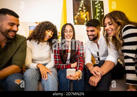 Group of multi ethnic young friends at home sitting on the sofa having fun looking at the smartphone - Caucasian woman with glasses and plaid shirt us Stock Photo