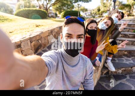 Outdoor group portrait of happy young people with medical mask - Millennial man take a selfie with his friends lined up behind him - New normal outdoo Stock Photo