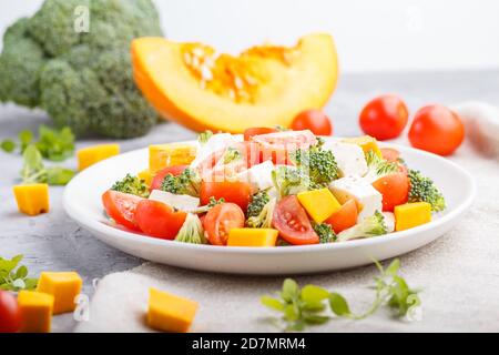 Vegetarian salad with broccoli, tomatoes, feta cheese, and pumpkin on white ceramic plate on a gray concrete background and linen textile, side view, Stock Photo