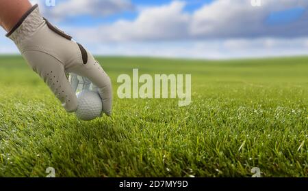 Golfer hand in a white glove holding a golfball, green course lawn background, close up view. Blue cloudy sky background. Stock Photo