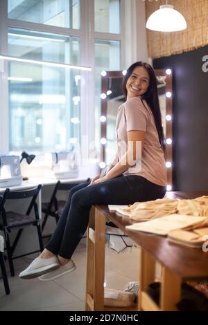 Happy young woman sitting on the pattern cutting table with fabric near sewing machines in workshop Stock Photo