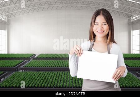 Happy female gardener with blank note in green house or glasshouse full of plants Stock Photo