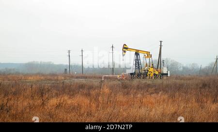 black and yellow metal pumpjack operates standing on yellow meadow under cloudy sky against white mist on winter day Stock Photo