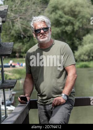 Handsome senior man with grey hair, beard, green T-shirt and sunglasses standing outdoors. Blurry background. Stock Photo