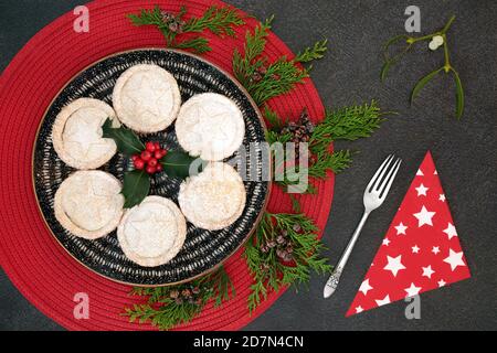 Christmas homemade mince pies with cedar cypress fir, winter berry holly, antique silver fork & napkin with mistletoe sprig on grey grunge background. Stock Photo