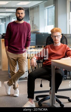 Two young serious businessmen in casualwear sitting and standing by workplace Stock Photo