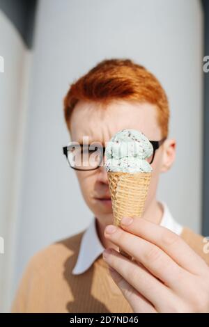 Serious young ginger man in glasses holding ice cream cone over his face Stock Photo