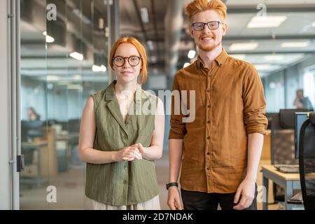 Young cheerful red haired colleagues in smart casualwear standing by each other Stock Photo