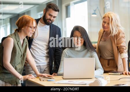 Pretty Asian businesswoman looking at laptop display among her happy colleagues Stock Photo