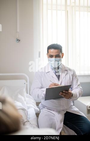 Young male doctor in formalwear, whitecoat and protective mask making notes Stock Photo