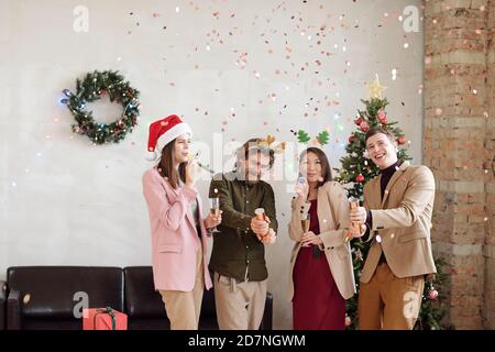 Joyful young colleagues in Christmas headwear shooting crackers and having fun Stock Photo