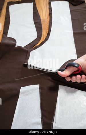 woman's hand cuts brown fabric by scissors according with pattern layouts of dress on wooden table at home Stock Photo