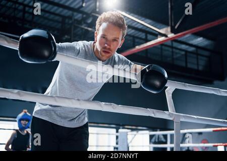 Tired young man in white shirt and protective gloves leaning on knots of boxing ring Stock Photo