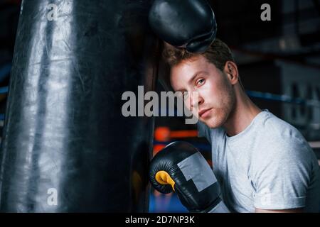 Tired young boxer in white shirt and with protective gloves leaning on punching bag in the gym Stock Photo