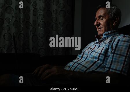 Side view of aged male remote worker in casual clothes using laptop while sitting in dark living room in late evening. Stock Photo