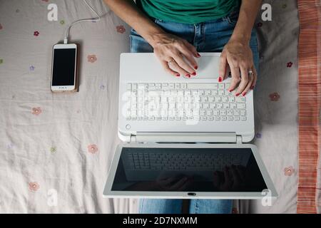 From above of crop anonymous female freelancer sitting on bed near smartphone and typing information on laptop keyboard while working remotely from ho Stock Photo