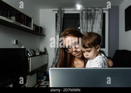 Serious mother holding toddler child while sitting at table and doing remote work on laptop at home. Stock Photo
