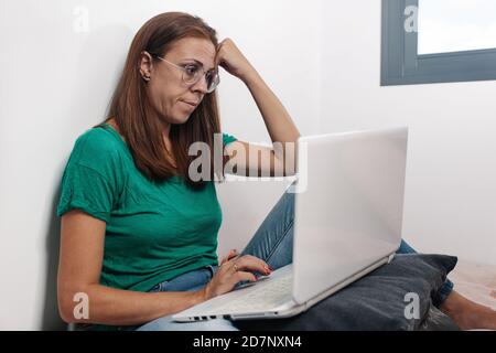 Concentrated middle aged female in casual wear and eyeglasses sitting on the bed and using laptop while working remotely from home Stock Photo