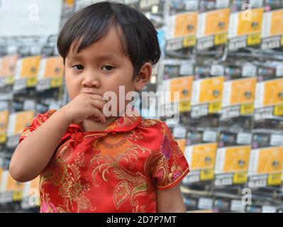 Dressed up cute little Thai Chinese girl wears a traditional red-and-gold Chinese silk dress (cheongsam) and sucks her thump during Chinese New Year. Stock Photo