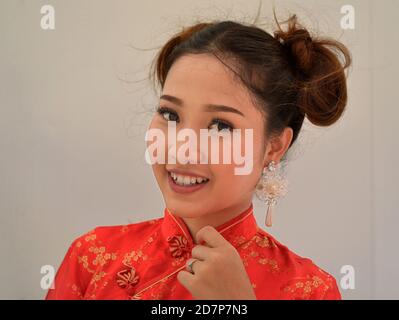 Beautiful young Thai Chinese woman wears a red-and-gold traditional Chinese silk dress (cheongsam) and smiles for the camera during Chinese New Year. Stock Photo