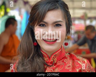 Young Thai woman with red dental braces, red lipstick and Chinese silk dress smiles for the camera inside a Buddhist temple during Chinese New Year. Stock Photo
