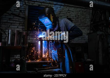 Side view of welder in protective helmet welding metal detail with sparks at factory Stock Photo