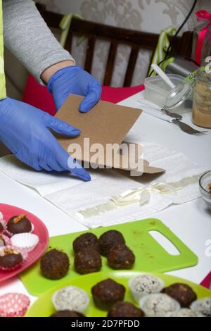 A woman in rubber gloves collects a cardboard box for sweets from dried fruits and nuts. Made at home in self-isolation during the epidemic. Stock Photo