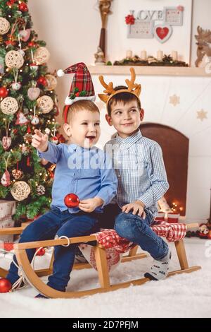 Cute and happy little brothers on sleigh having fun in front of Christmas tree and fireplace - Merry Christmas Stock Photo