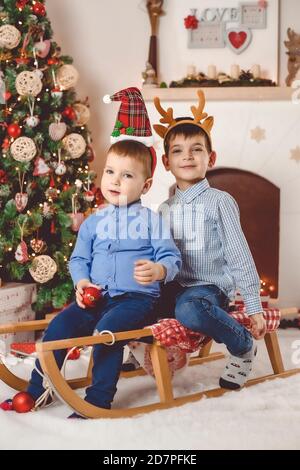 Cute and happy little brothers on sleigh having fun in front of Christmas tree and fireplace - Merry Christmas Stock Photo