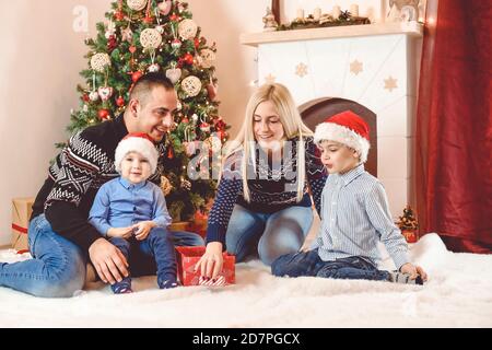 Mother, father and kids celebrate Christmas - they sitting in front of Christmas tree, playing and opening gifts Stock Photo