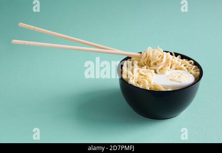 Cooked instant noodles with egg in black plastic bowl with chopsticks on a blue paper background with place for text. Chinese chopsticks float in the Stock Photo