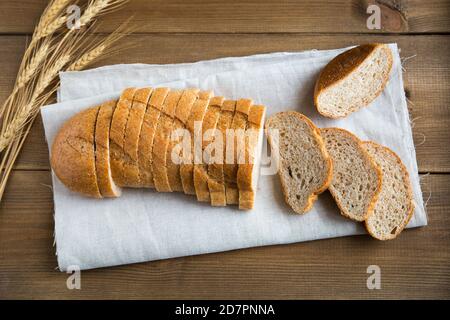 A loaf of wheat bread with bran is cut into slices and lies on a linen napkin. Several whole, dry ears of wheat lie nearby. Flat lay, horizontal, clos Stock Photo
