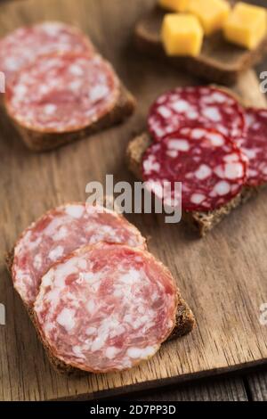 Sandwiches with smoked salchichon and braunschweig sausages are on wooden board. Next to are cheese cubes on bread. Close up, vertical composition. Stock Photo