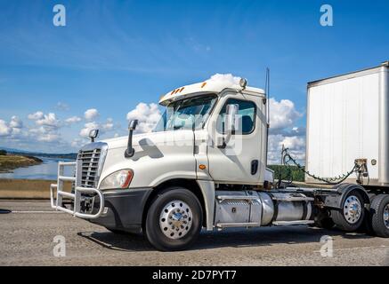 Powerful big rig white industrial grade day cab diesel semi truck with pipe grille guard transporting commercial cargo in dry van semi trailer running Stock Photo
