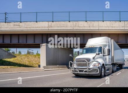 Powerful big rig white industrial grade day cab diesel semi truck with pipe grille guard transporting commercial cargo in dry van semi trailer running Stock Photo