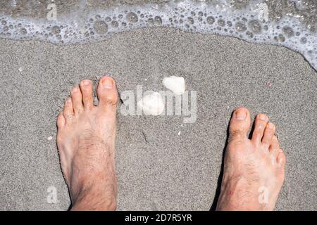 Male feet closeup point of view standing by white seashells sea shells during shelling fun activity on Sanibel Island, Florida USA on shore wave water Stock Photo