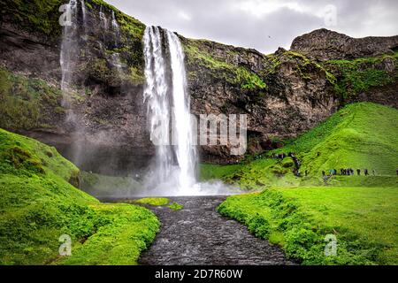 Seljalandsfoss waterfall in Iceland with white water falling off cliff in green lush summer landscape and river with people and cloudy sky Stock Photo