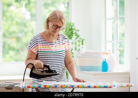 Senior woman ironing clothes. Female folding clothes at iron board. Home chores. Housewife cleaning house. Stock Photo
