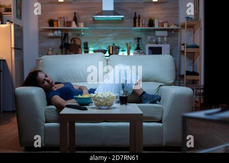 Young woman watching TV and feeling bored sitting on the couch in the living room at home. Tired, exhausted, lonely lady relaxing watching tv lying on comfortable sofa eating snacks late at night Stock Photo