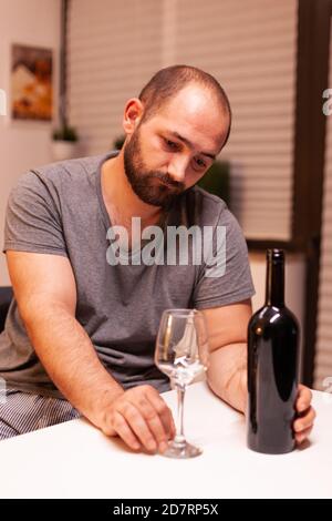 Man in dispair drinking alcohol alone sitting at table in kitchen. Unhappy person disease and anxiety feeling exhausted with dizziness symptoms having alcoholism problems. Stock Photo