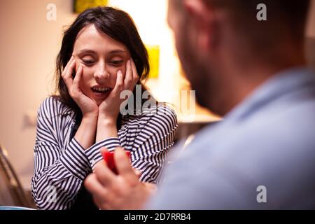 Woman looking happy at engagement ring during marriage proposal while having romantic dinner. Man asking his girlfriend to marry in the kitchen during romantic dinner. Happy caucasian woman smiling being speechless Stock Photo