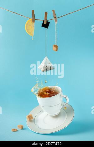 Tea bag, lemon and sugar hanging on the rope over splashing tea on blue background. Creative still life concept. Stock Photo