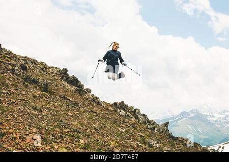 Happy young female tourist jumps on top of a mountain with climbing sticks in her hands against the background of a high snowy mountain while travelin Stock Photo