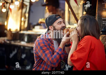 cute caucasian couple eating one sandwich together, taking bite each from another side of it Stock Photo