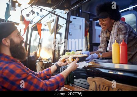 polite afro-american female employee holding, giving, two sandwiches to a satisfied beardy caucasian customer