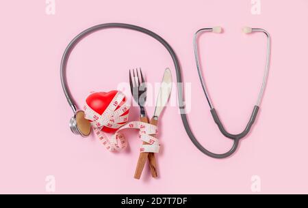 Red heart and stethoscope with measuring tape around fork and knife isolated on pink background, health care and diet concept Stock Photo