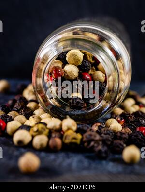 Close-up of 4 kinds of peppercorns spilling out ot a glass bottle on a dark textured surface Stock Photo