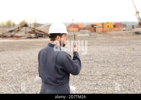 Back view of foreman standing on construction site, talking by VHF walkie talkie and holding blueprints. Stock Photo