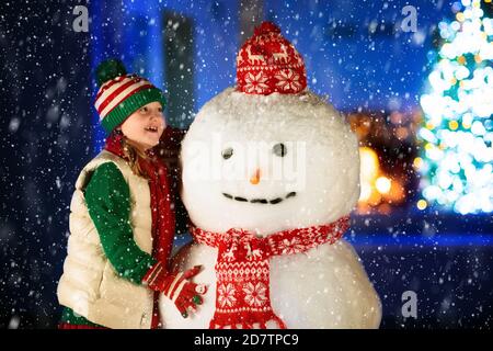 Kids build snowman on Christmas eve. Child with presents and gifts in snowy backyard next to window to living room with Christmas tree and decorated f Stock Photo