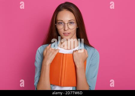 Close up portrait of young woman with glasses holding text book organaiser, looking at camera, wearing round spectacles, reads interesting book, havin Stock Photo